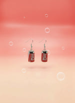 Coca-Cola can-shaped earrings on a black card with a wooden stand against a white background