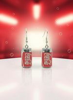 Coca-Cola can-shaped earrings on a black card with a wooden stand.