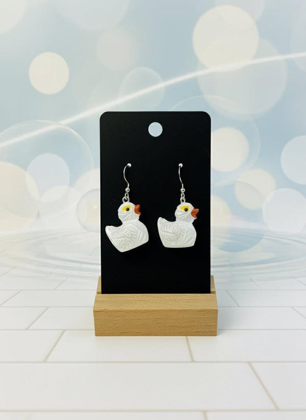 White duck-shaped earrings on a black card with a wooden stand against a white background
